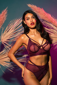 Harness style bra with mesh overlay and visible gold hardware in a deep wine red/purple, worn with a matching brief in front of a tropical backdrop.