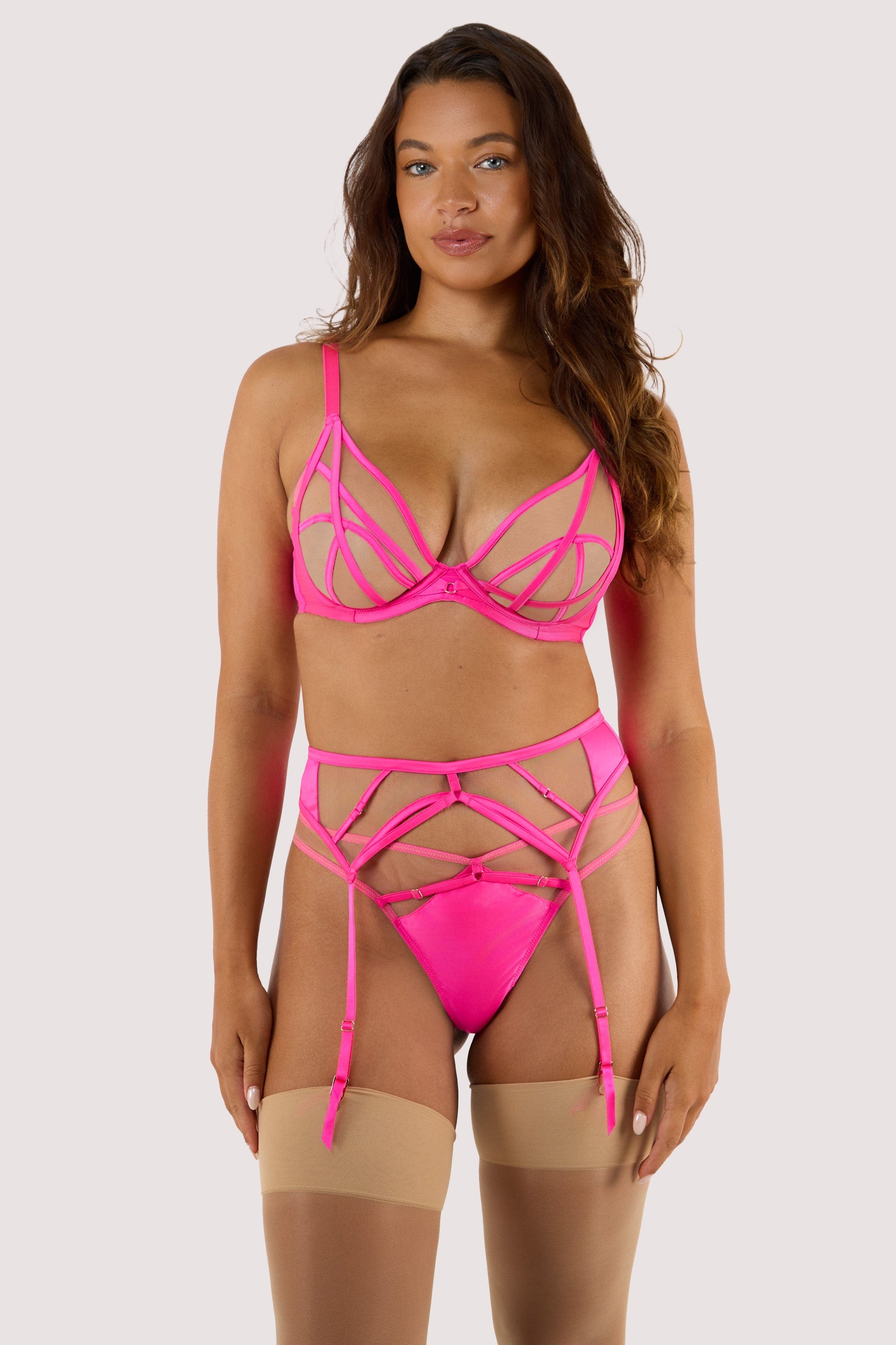 model wears neon pink illusion mesh suspender belt with satin strap cage detailing, sheer panels, and gold hardware, styled with matching bra, thong, and nude stockings